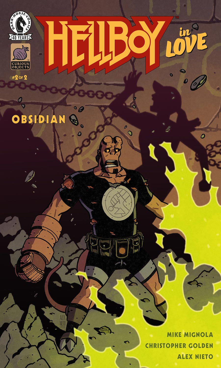 Comic book cover for Hellboy in Love: Obsidian, issue #2, published by New Zealand Comics. The cover art features Hellboy in a black and brown outfit with a circular chest emblem, standing in a dark, rocky environment with a shadowy figure and bright yellow-green energy effects.